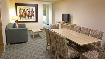 A hotel room with a dining area featuring a wooden table and patterned chairs, a sofa with cushions, a television on a stand, and a framed artwork on the wall.