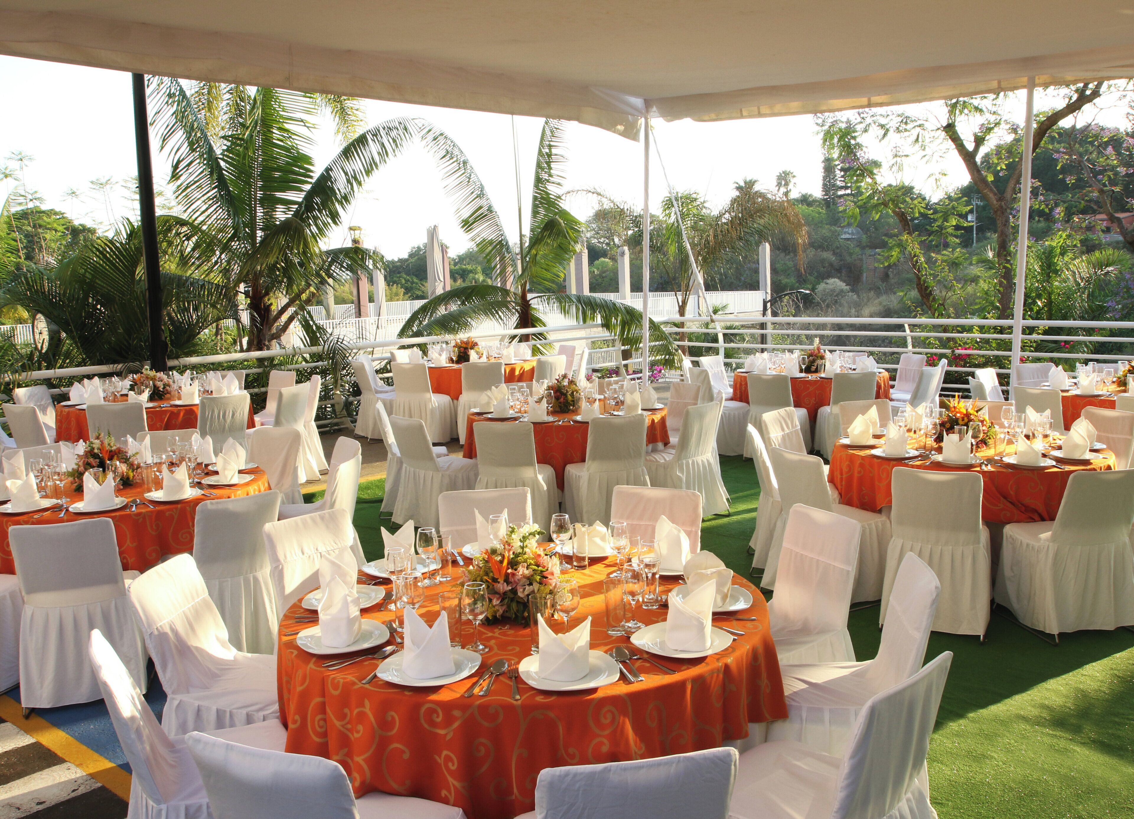 An outdoor dining area with round tables, white chairs, and orange tablecloths.