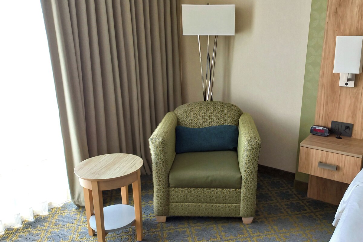 A hotel room with a green patterned armchair, a small wooden table, a bedside table with a lamp, and a bed with white linens.