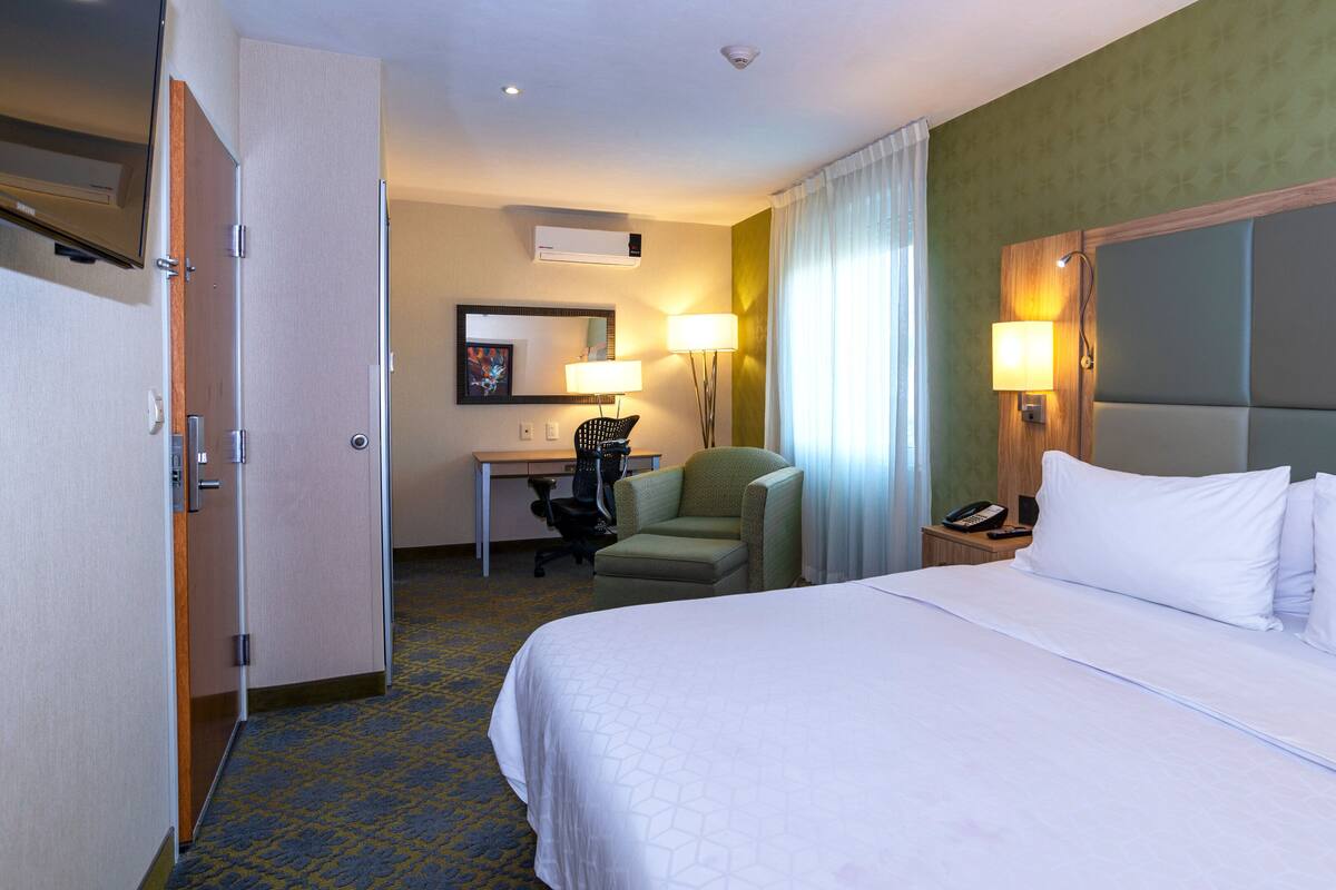 A hotel room with a bed, desk, chair, and a small table with a lamp.
