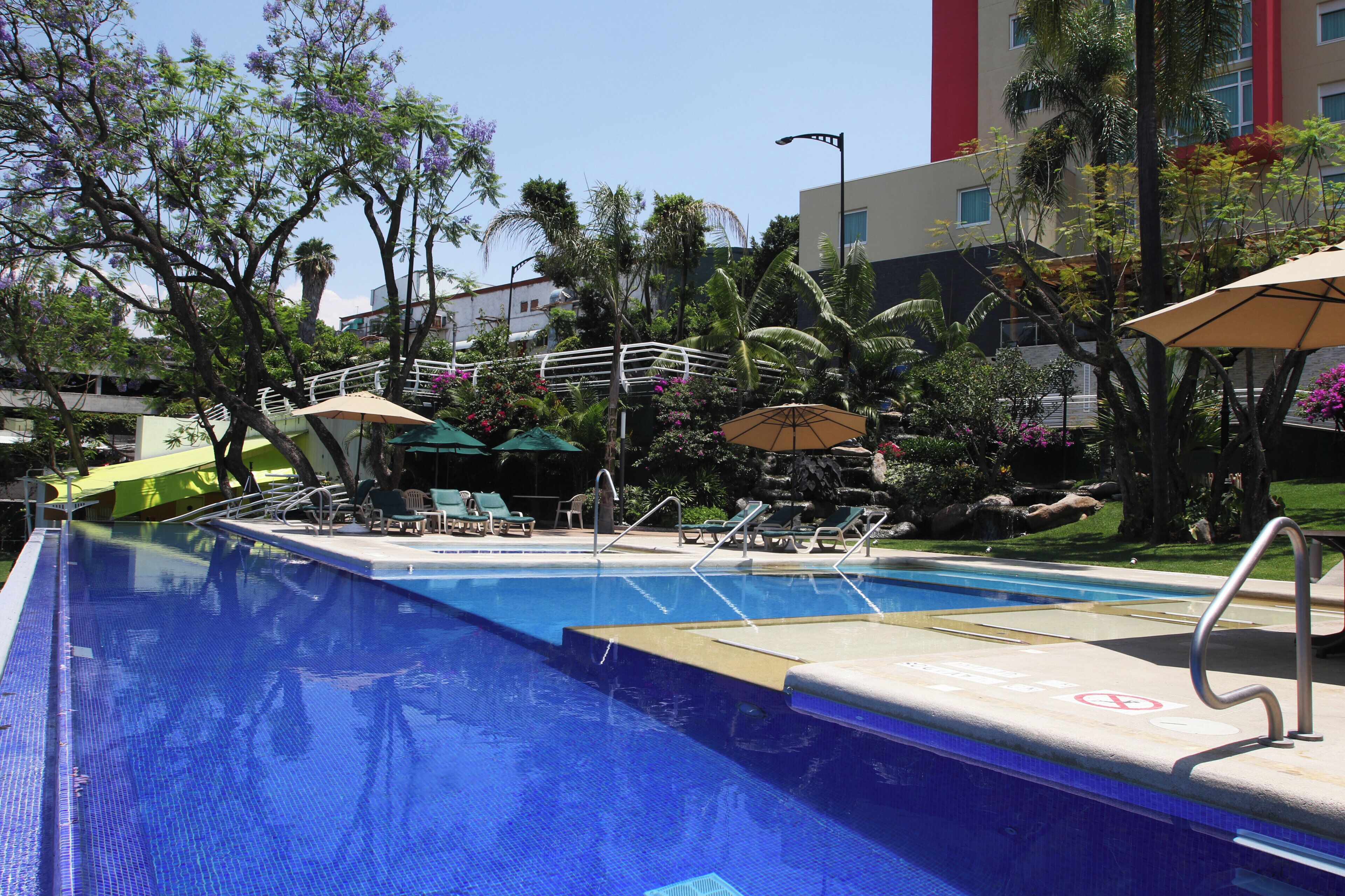 Una piscina con sillas de descanso y sombrillas, rodeada de palmeras y un edificio con fachada roja y blanca.