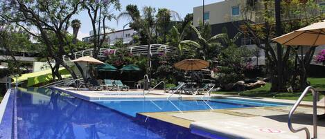 Una piscina con sillas de descanso y sombrillas, rodeada de palmeras y un edificio con fachada roja y blanca.