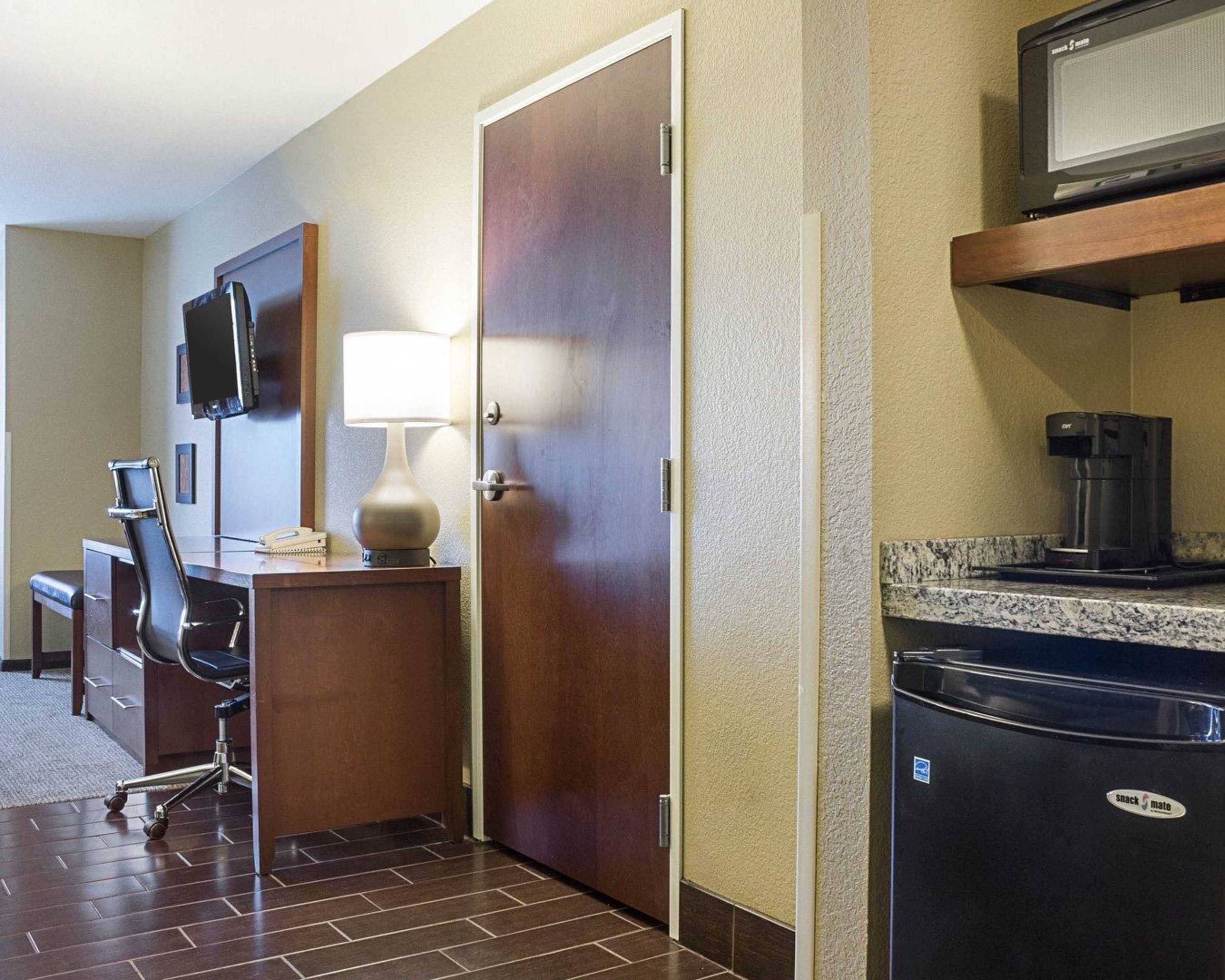 A hotel room with a desk, chair, lamp, television, microwave, and refrigerator.