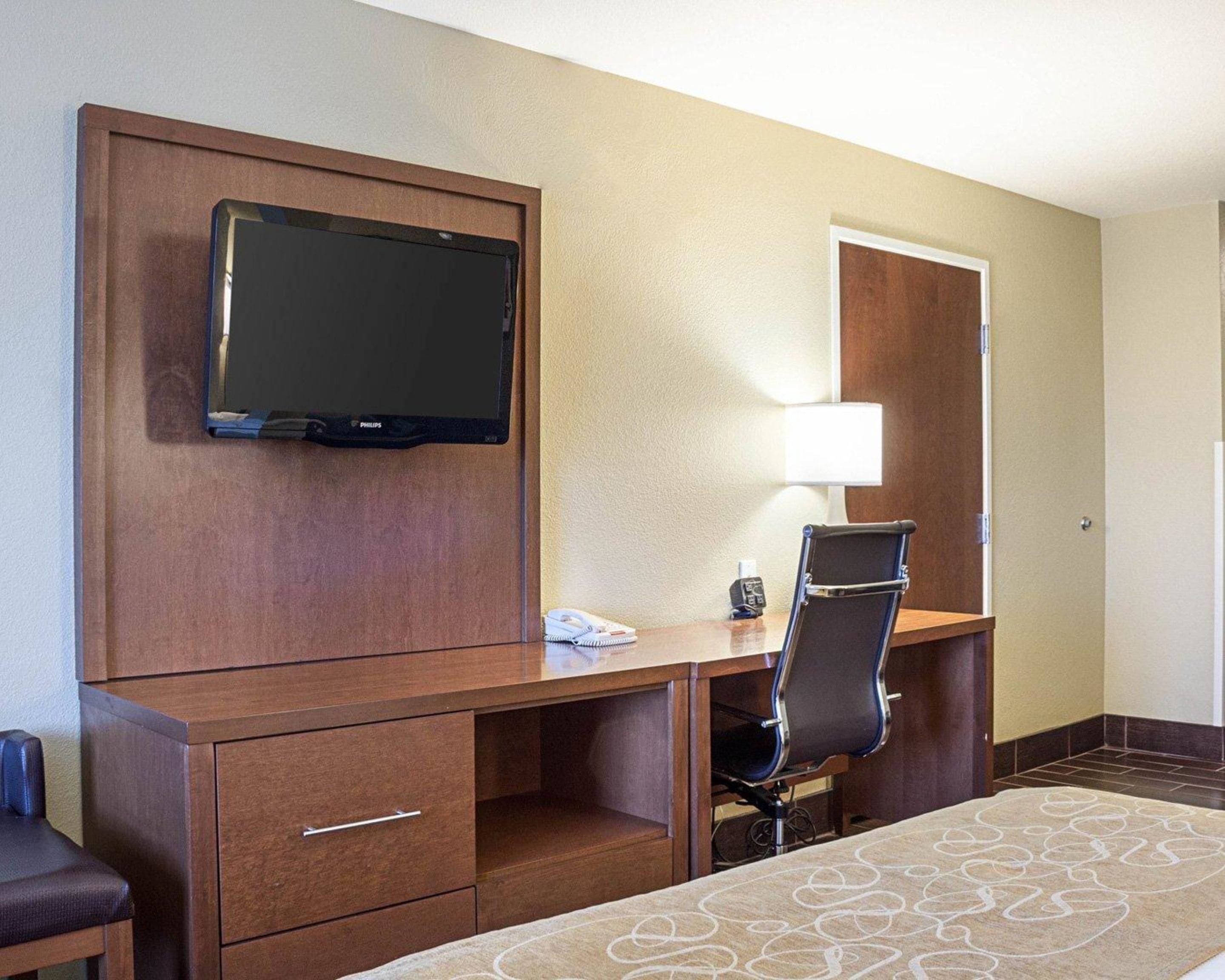 A hotel room with a wooden desk, a flat-screen TV, a chair, a lamp, and a bed with a patterned bedspread.