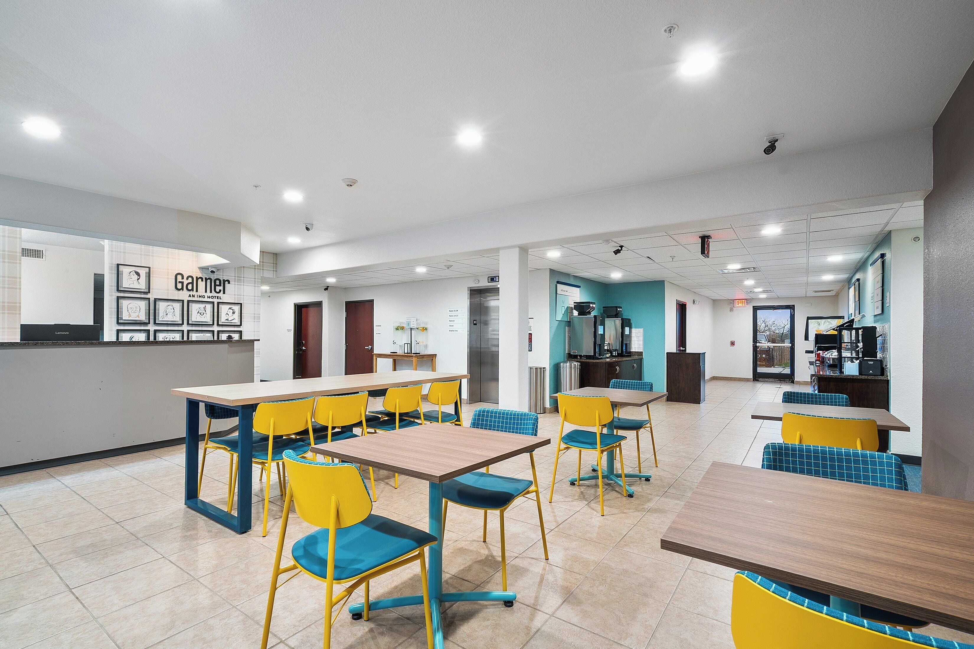 A hotel dining area with multiple tables and chairs, a kitchen in the background, and a view of the outside.