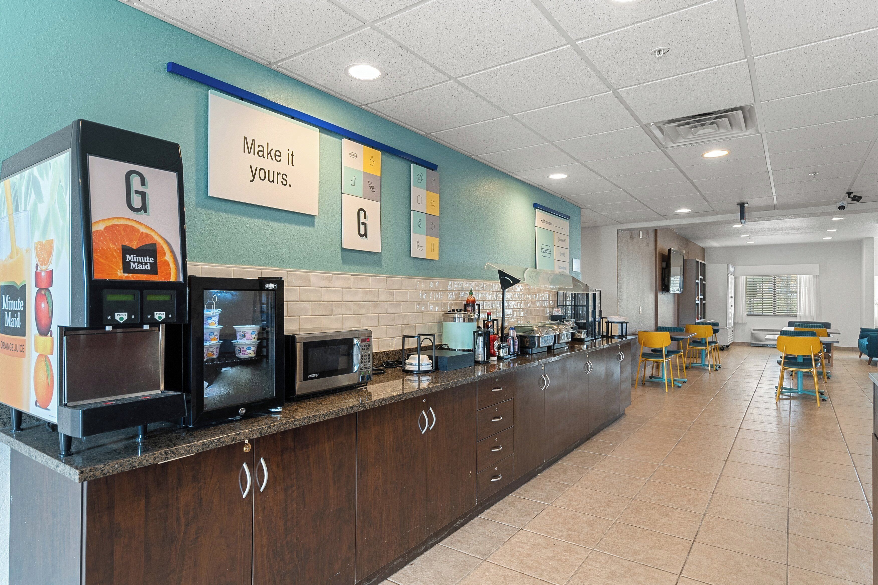 A clean, well-lit food service area with a counter, microwave, and coffee machines. There are signs on the wall and a seating area with tables and chairs.