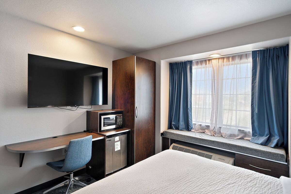 A hotel room with a bed, a desk with a chair, a microwave, a TV, and a window with curtains.
