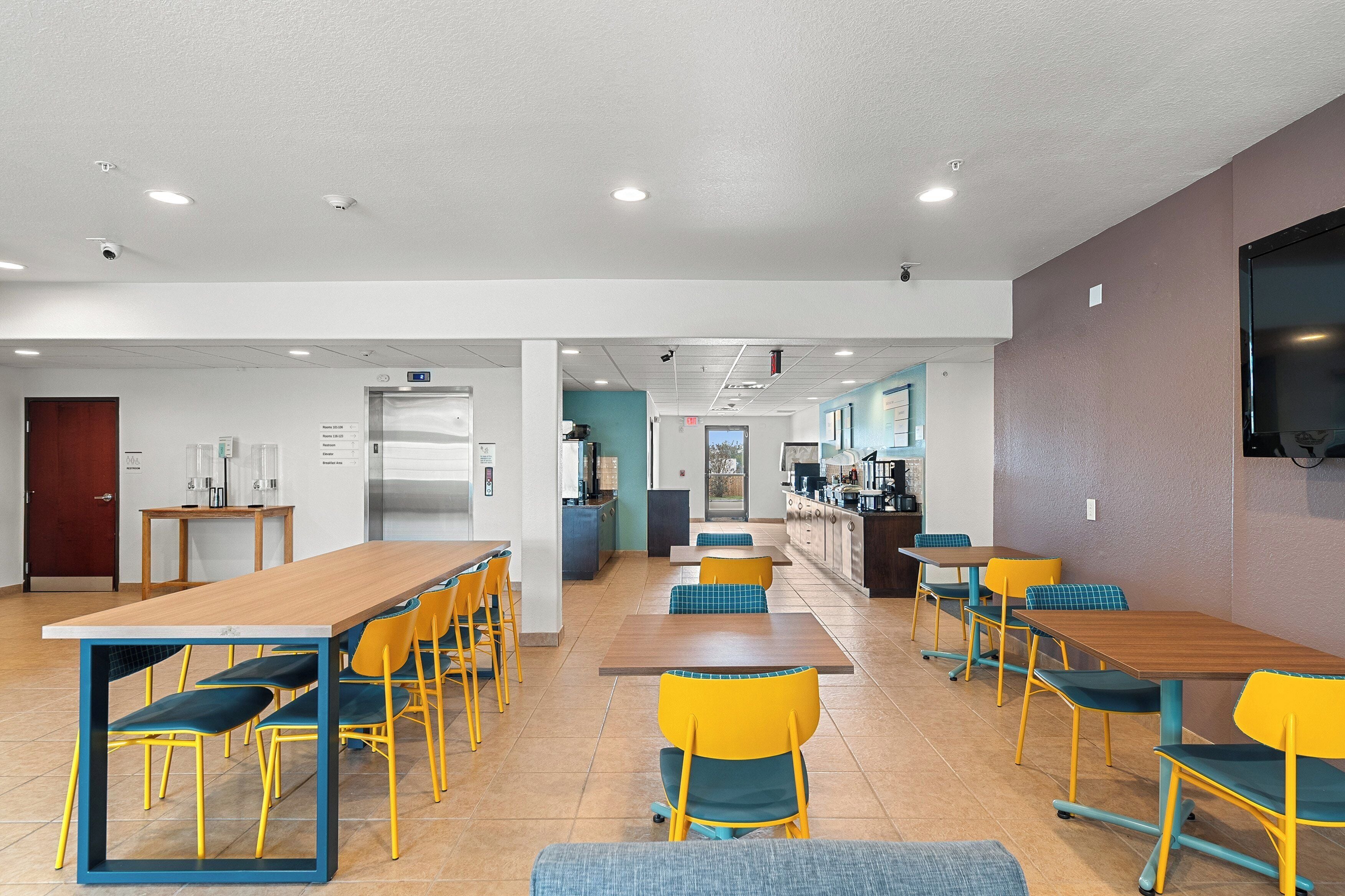 A spacious dining area with multiple tables and chairs, a kitchen area in the background, and a large screen on the wall.
