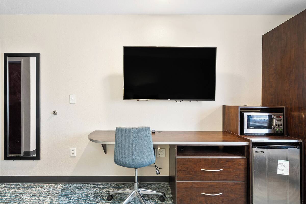 A hotel room with a desk, chair, flat-screen TV, microwave, refrigerator, and a mirror.