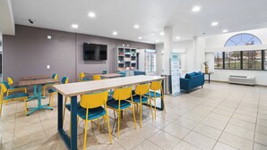 A spacious indoor area with a long table and yellow chairs, a blue sofa, a TV mounted on the wall, and vending machines. - Garner Hotel Gonzales by IHG (Gonzales)