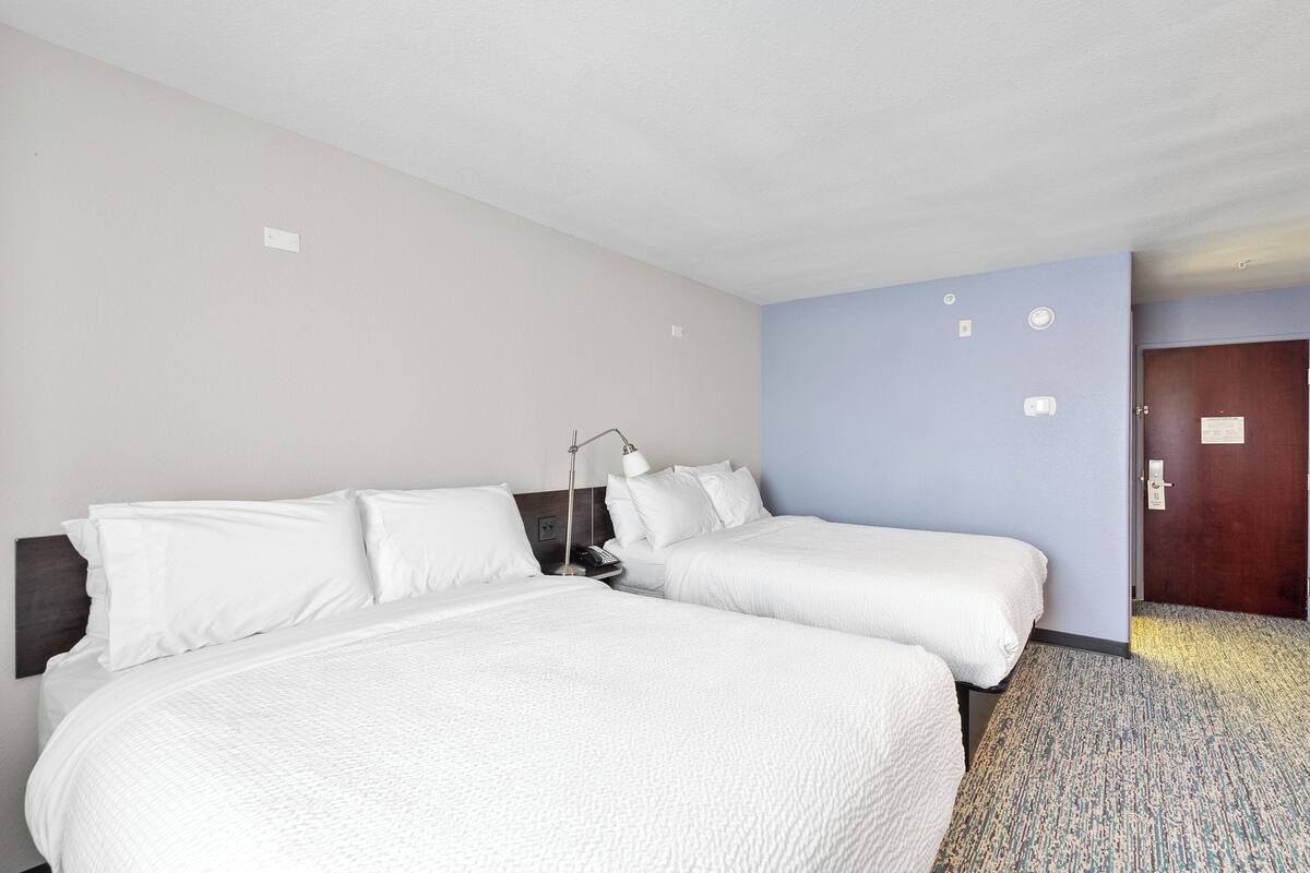 A hotel room with two beds, a nightstand, a lamp, and a door to another room.