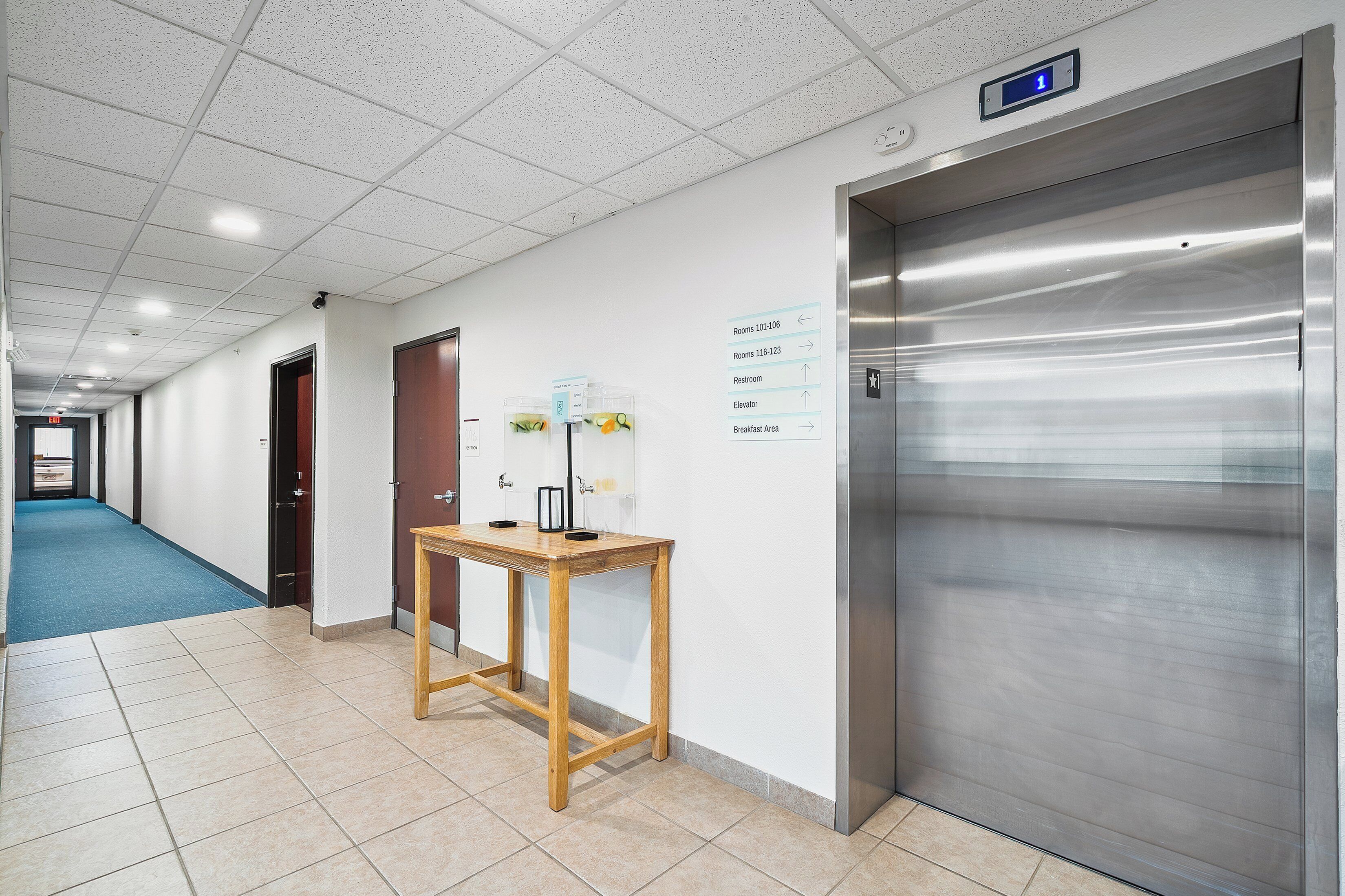Pasillo de hotel con ascensor, un cartel con los números de las habitaciones y una mesa con dispensador de agua.