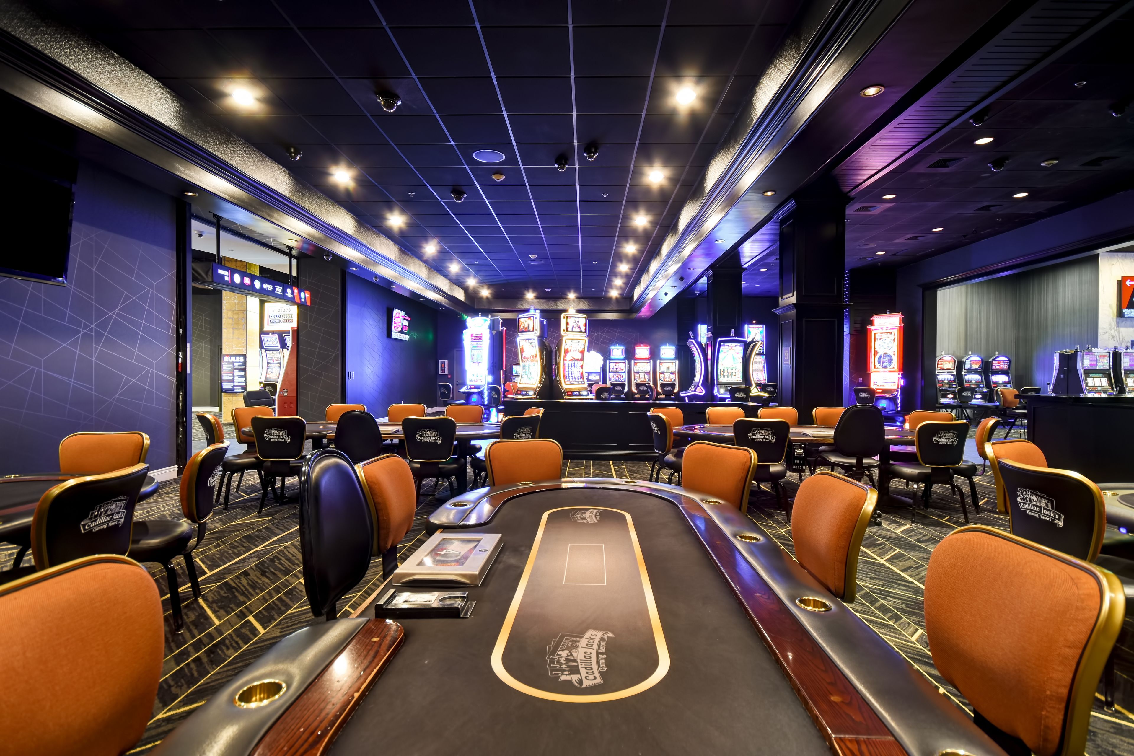 Een casino met meerdere blackjack tafels, oranje en zwarte stoelen, en gokkasten.