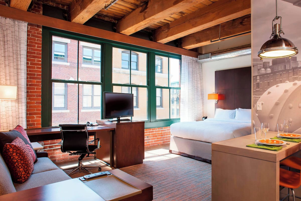 A hotel room with a brick wall, large windows, a bed, a desk with a chair, a sofa, and a dining area with plates and glasses.