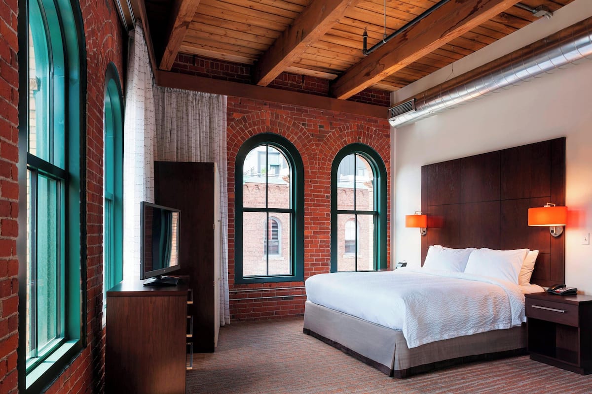 A hotel room with a brick wall, large arched windows, a wooden ceiling, a bed, a nightstand, and a TV on a cabinet.