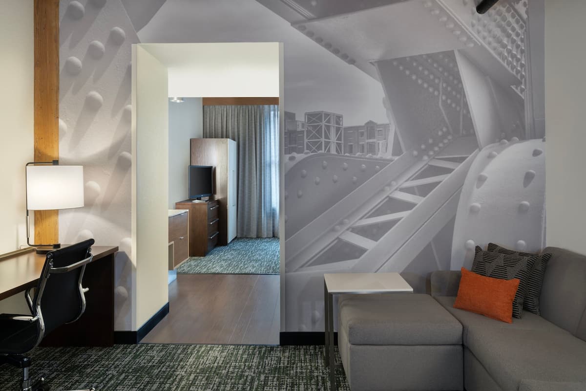 A hotel room with a modern design, featuring a desk, a sofa, and a view of a cityscape through a large window.