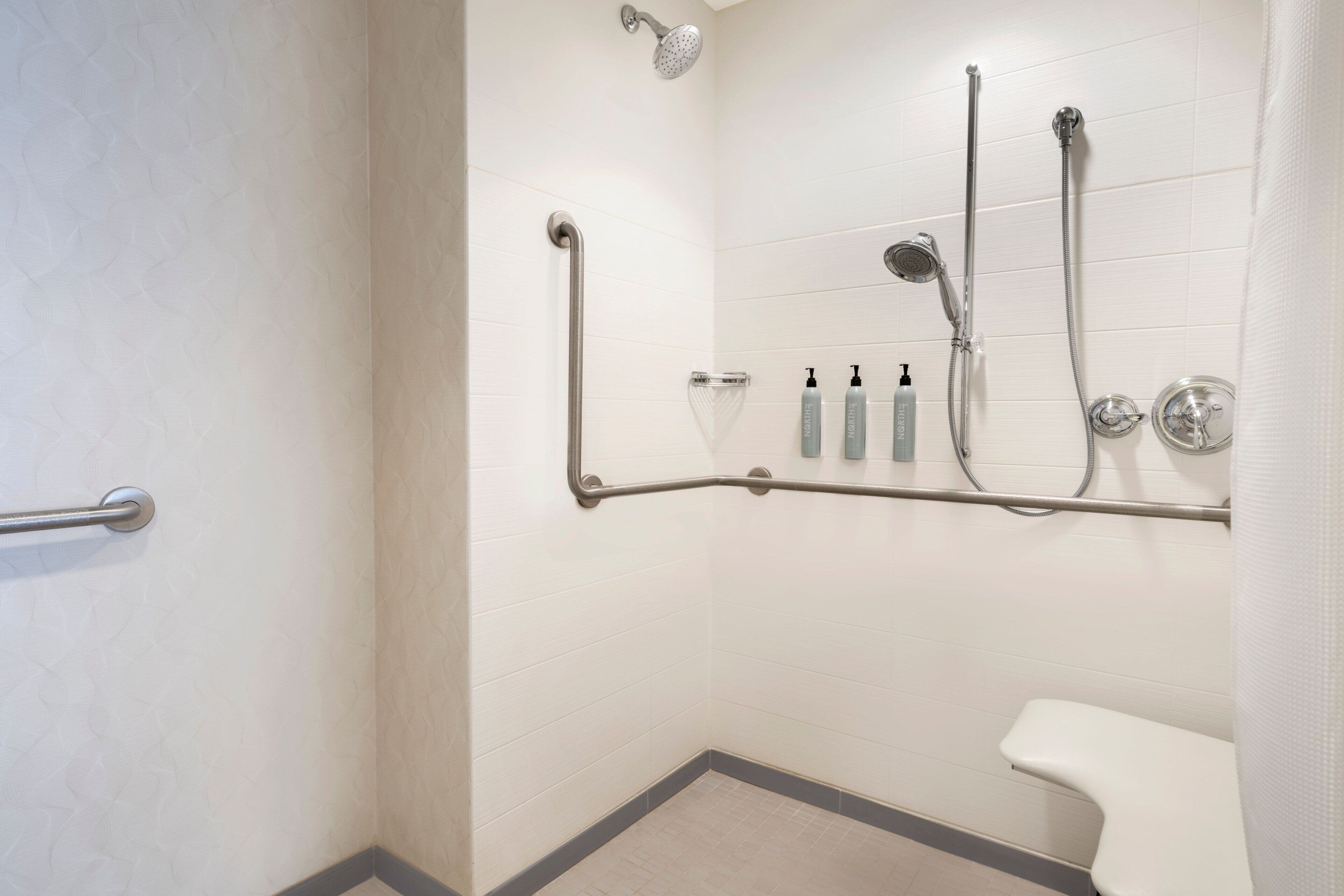 Un baño con inodoro blanco, ducha con cabezal de ducha de mano y barra de apoyo.