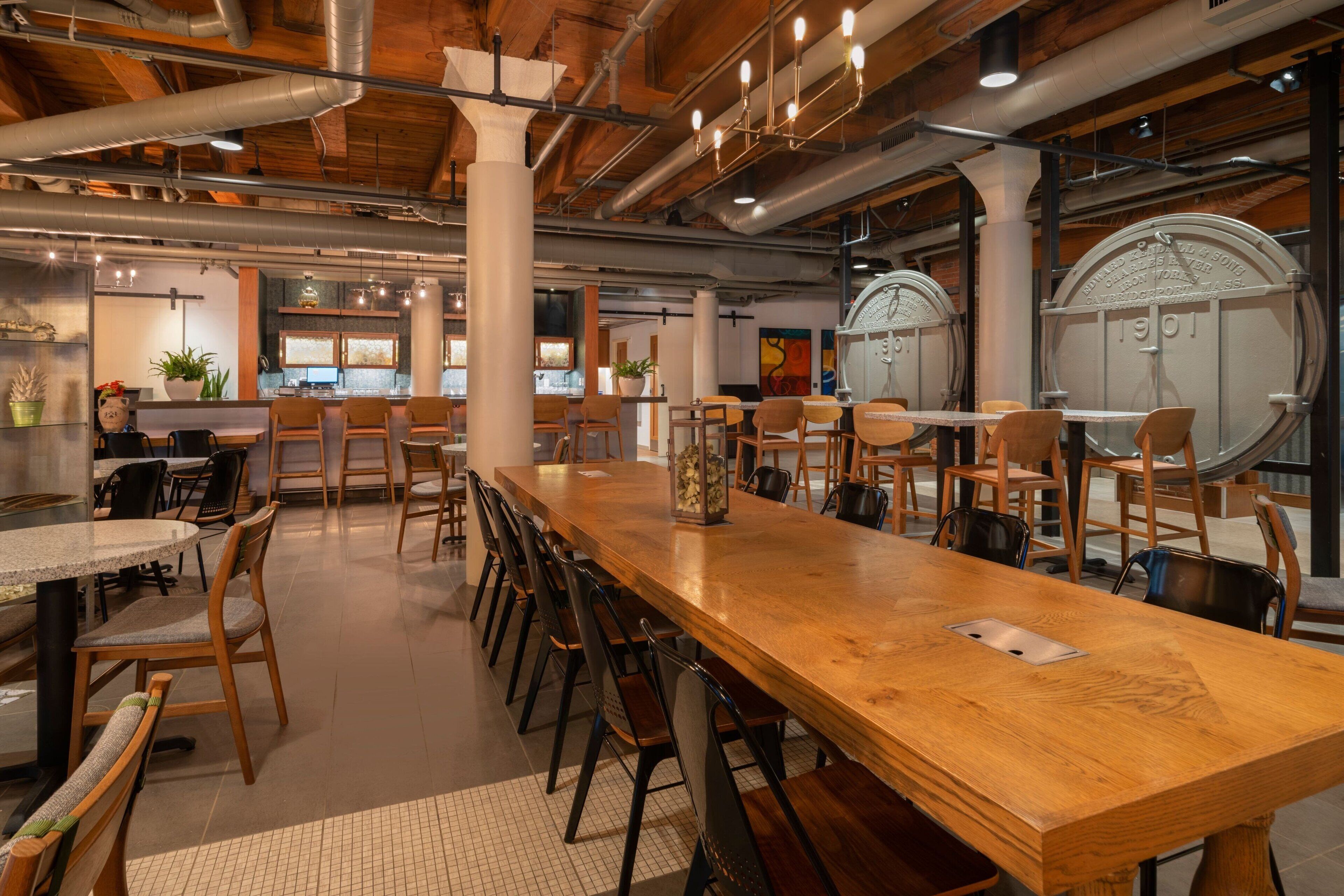 A spacious dining area with a long wooden table and multiple chairs, surrounded by high ceilings with exposed ductwork and wooden beams.