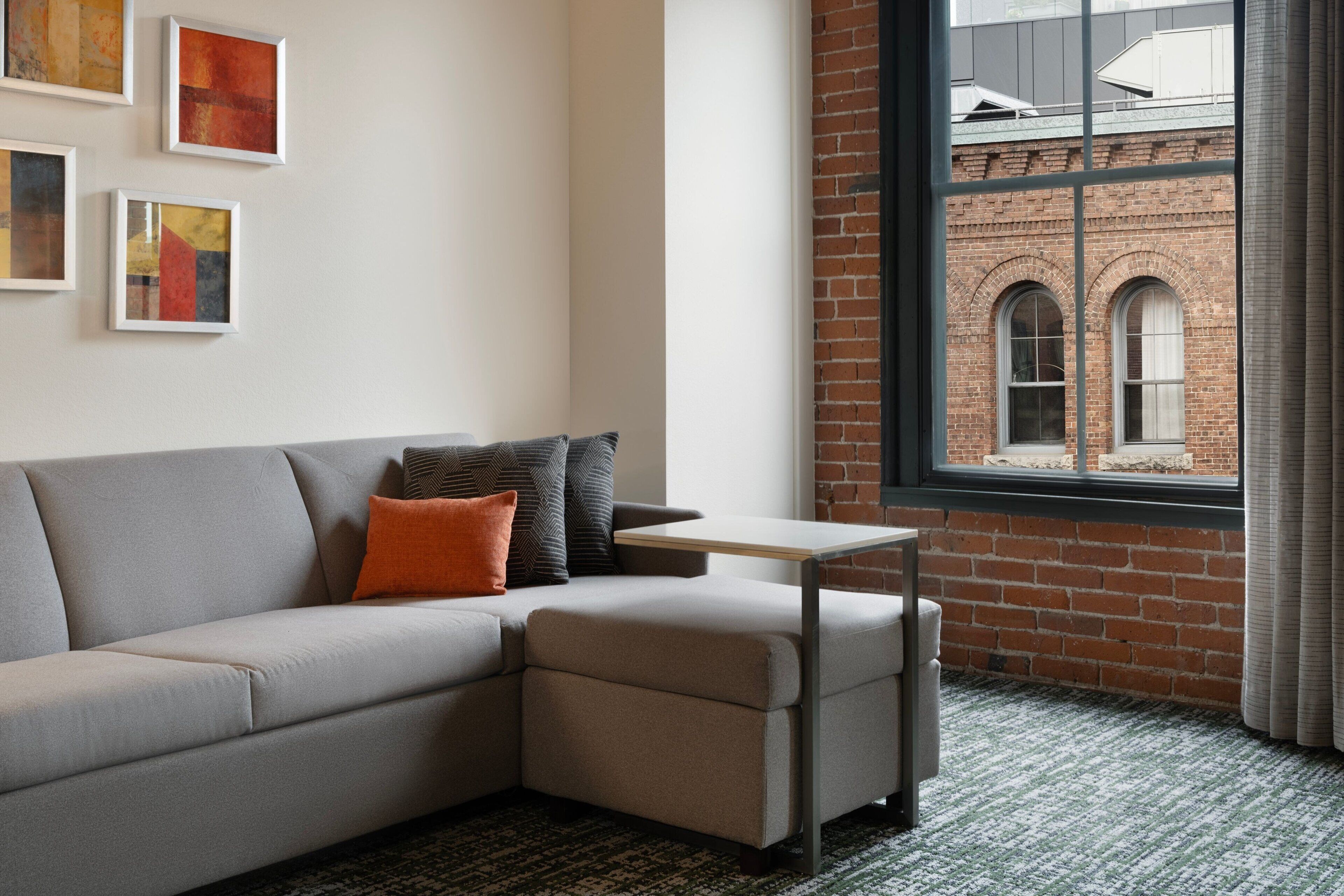 A modern living room with a grey sectional sofa, a small table, and a view of a brick building through a large window.