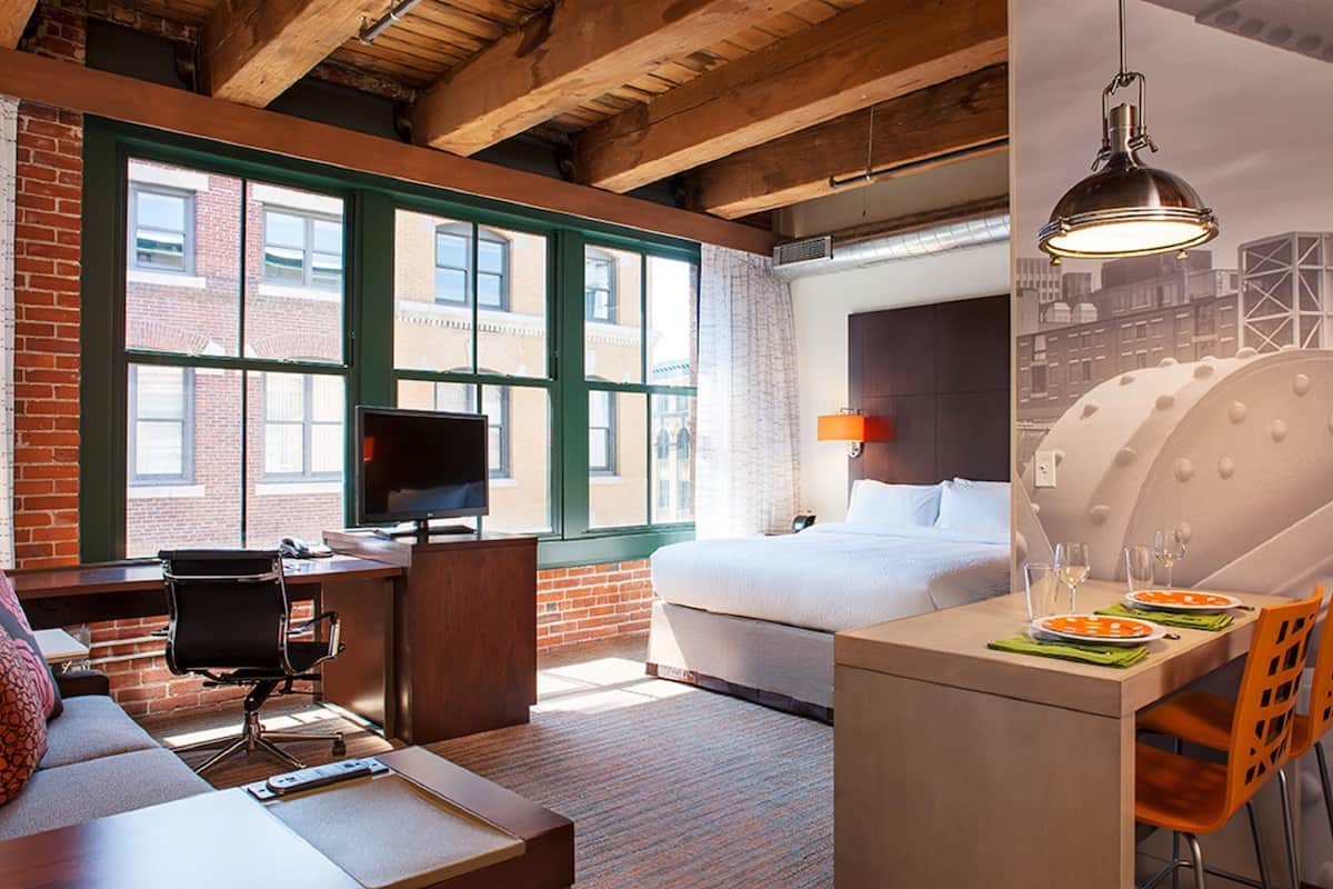 A hotel room with a brick wall, large windows, a bed, a desk with a computer, a sofa, and a dining area with chairs and a table.
