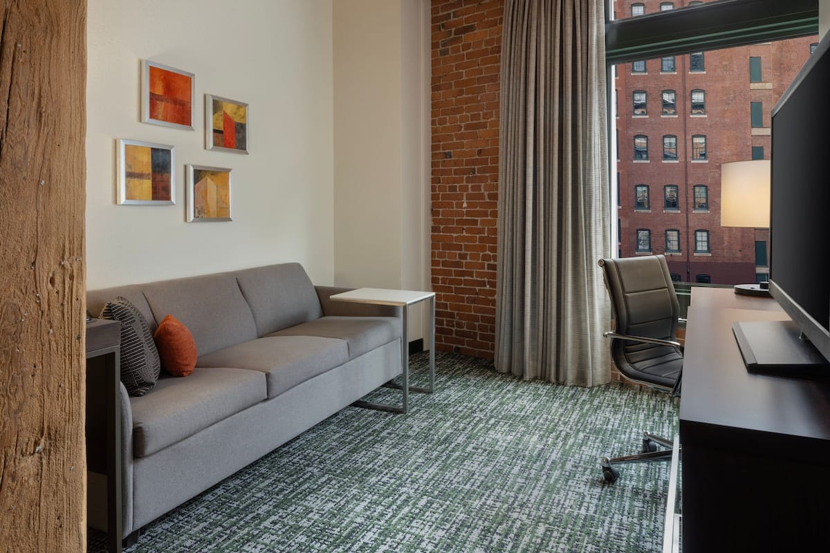 A hotel room with a grey sofa, a small table, a chair, a TV, and a window with a view of a brick building.