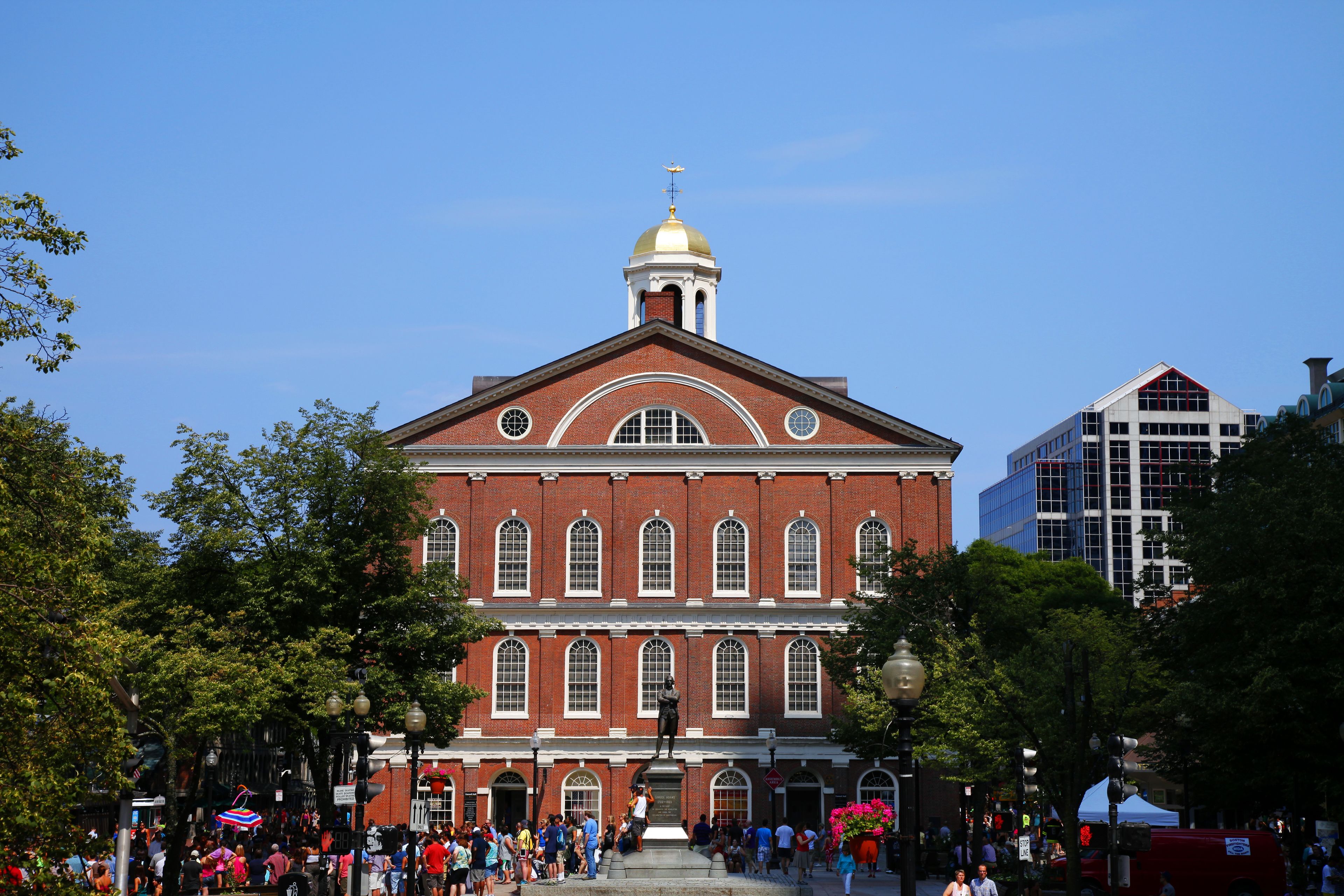 A historic building with a central dome and arched windows, surrounded by trees and a crowd of people.