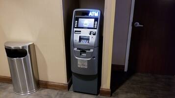 An ATM machine with a trash can beside it, located in a hallway with a wooden door.