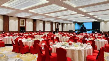 A large banquet hall with round tables covered in white tablecloths, red chairs, and a high ceiling with a decorative light fixture.