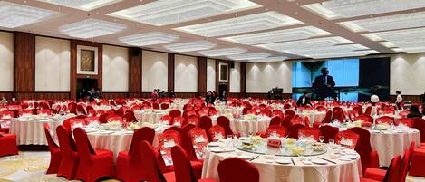 Une vaste salle de banquet avec des tables rondes recouvertes de nappes blanches, des chaises rouges et un plafond haut orné d’un luminaire décoratif.
