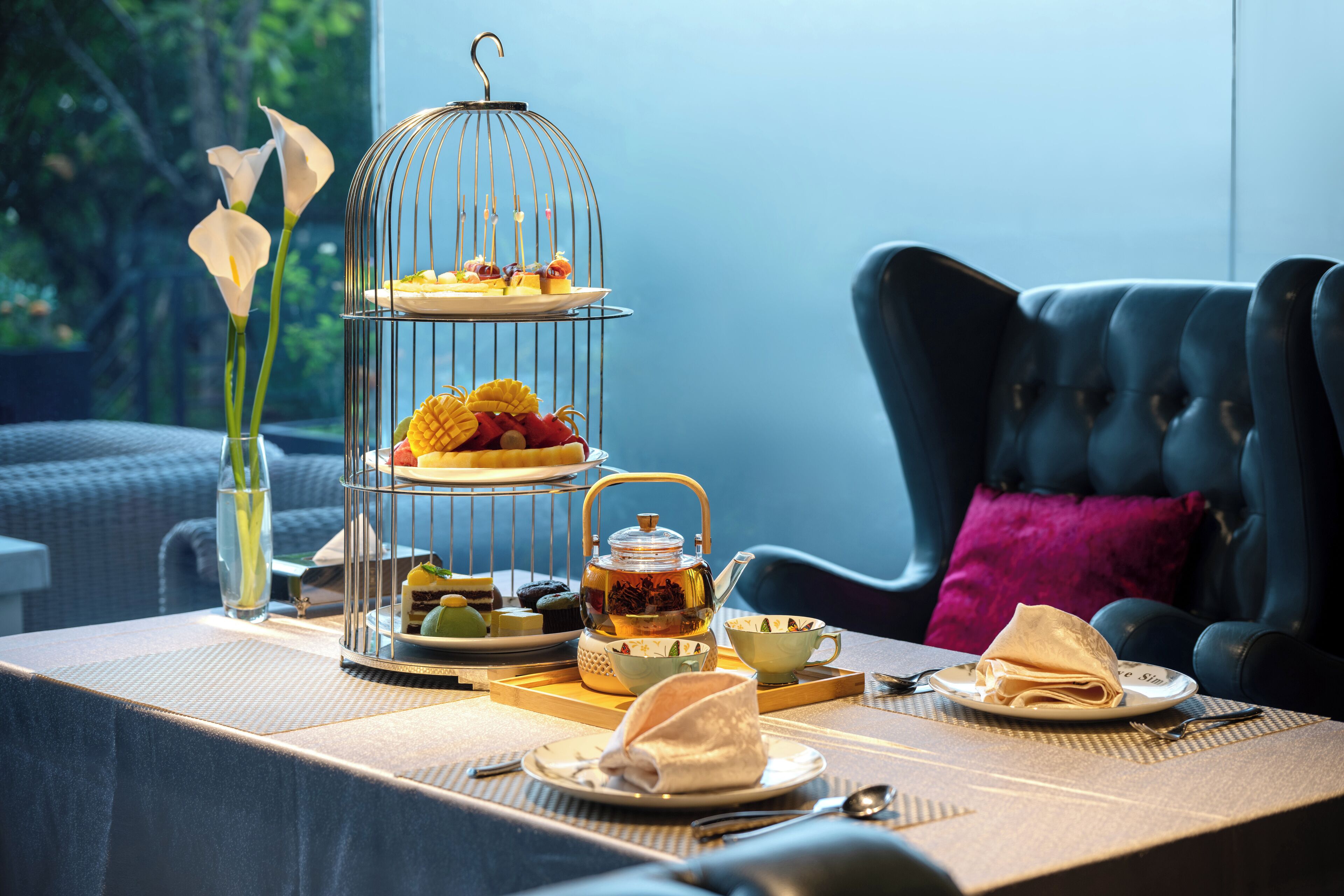 Una mesa de comedor dispuesta con un soporte escalonado para postres, una tetera y un florero con flores.