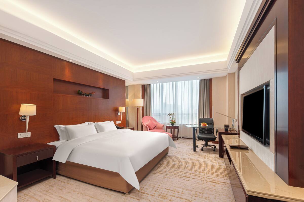 Ein Hotelzimmer mit einem großen Bett, einem Schreibtisch mit Flachbildfernseher, einem Sessel, einem kleinen Tisch und einer Lampe.