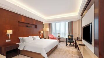 A hotel room with a large bed, a desk with a flat-screen TV, a chair, a small table, and a lamp.