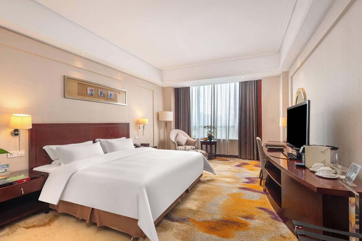Ein Hotelzimmer mit einem großen Bett, einem Schreibtisch mit Computer, einem Fernseher und einem Sessel.