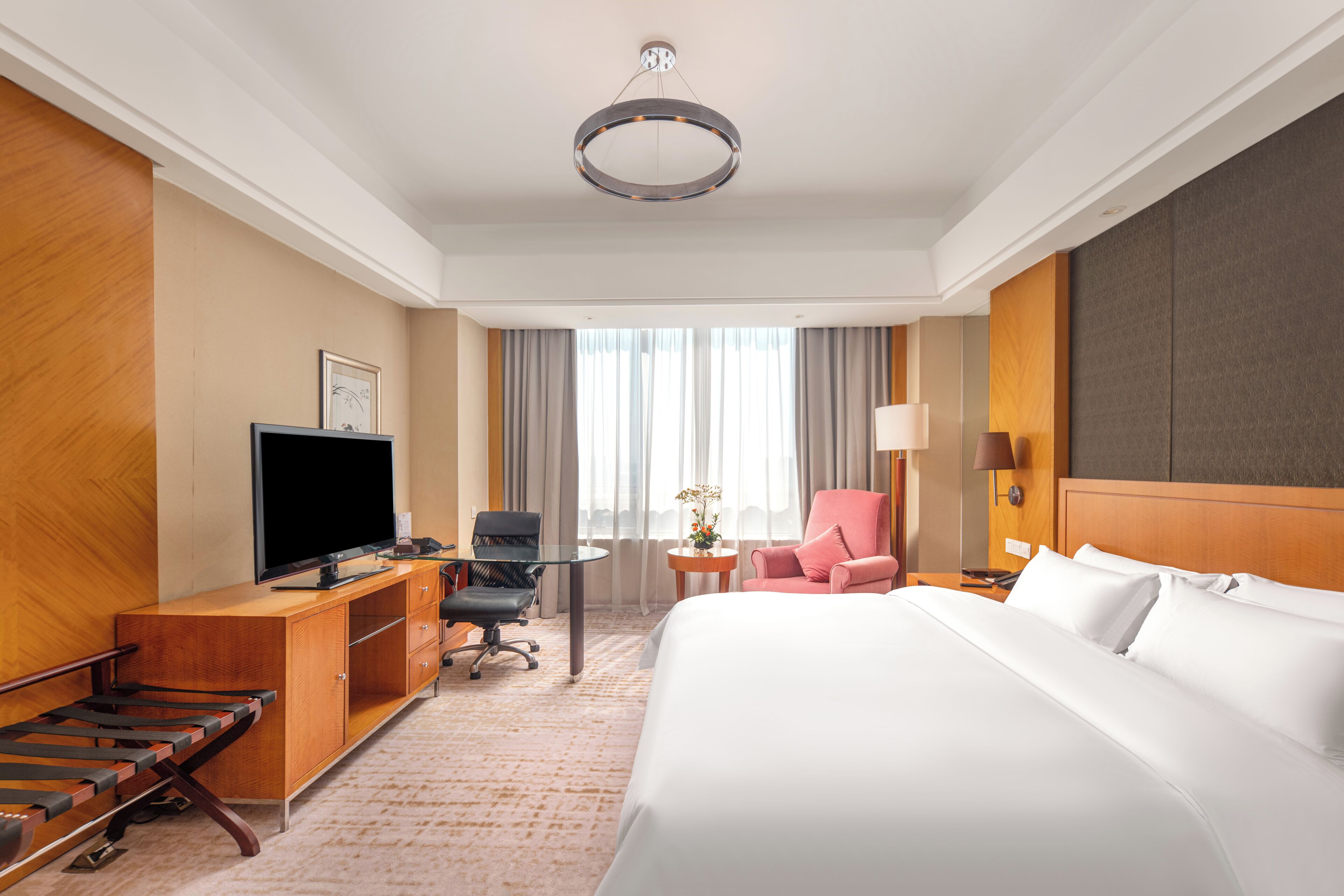 Habitación de hotel con una cama, un escritorio con televisor, una silla, una lámpara y una ventana con cortinas.