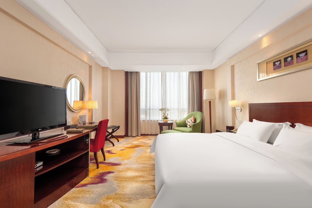 Ein Hotelzimmer mit einem großen Bett, einem Fernseher, einem Schreibtisch mit Stuhl und einem fensterbepflanzten Fenster.