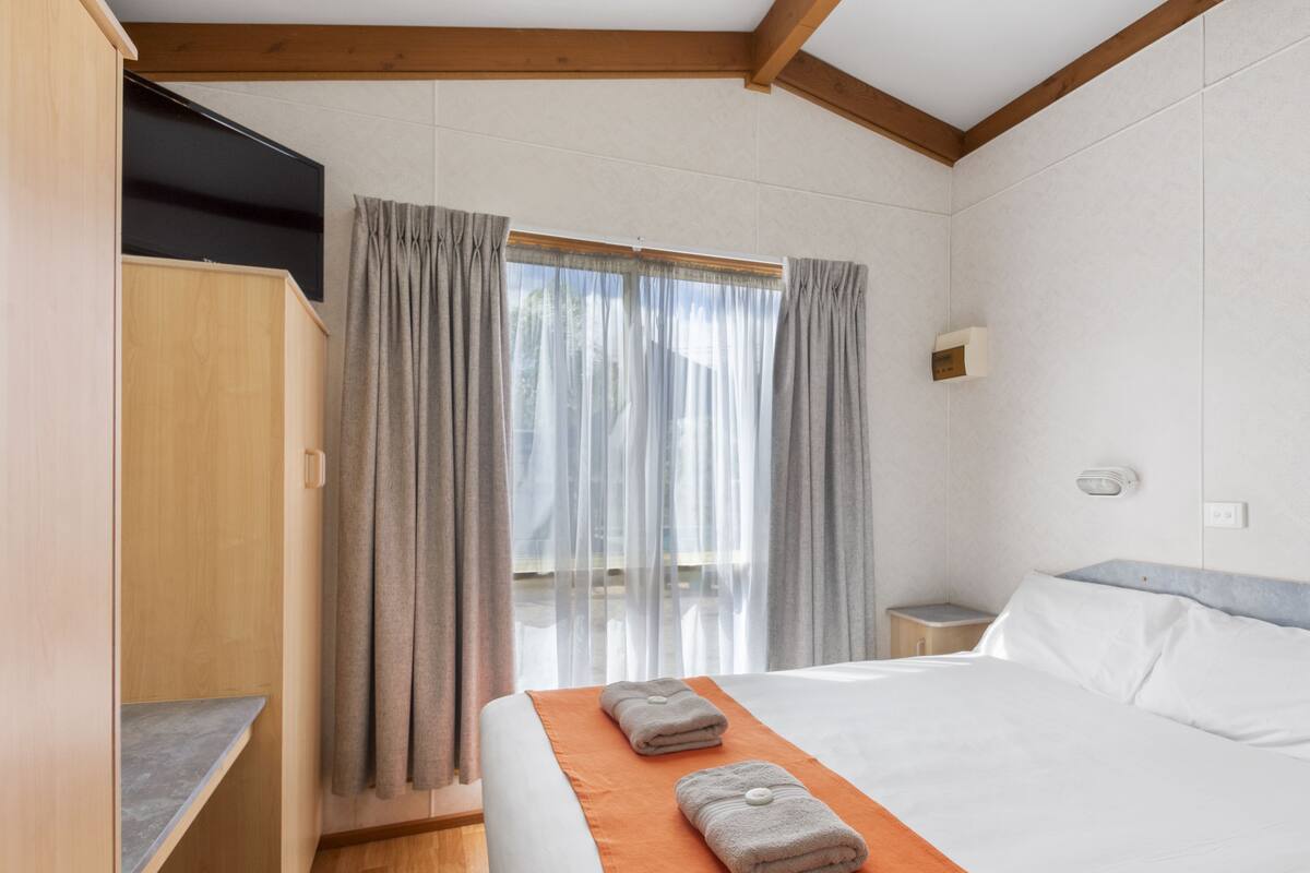 Una habitación de hotel con cama, armario de madera, ventana con cortinas y mesita de noche.