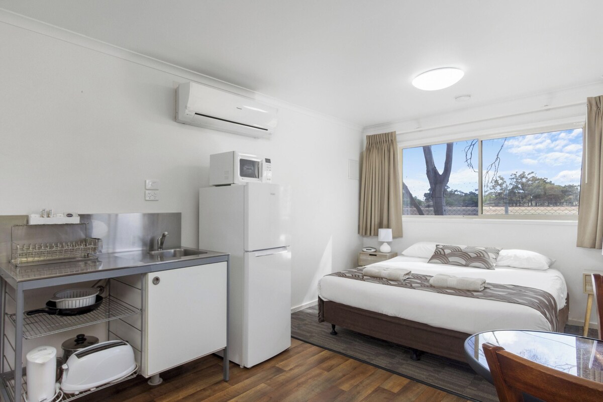 A compact hotel room with a kitchenette, a bed with patterned bedding, a window with a view, and a small table.