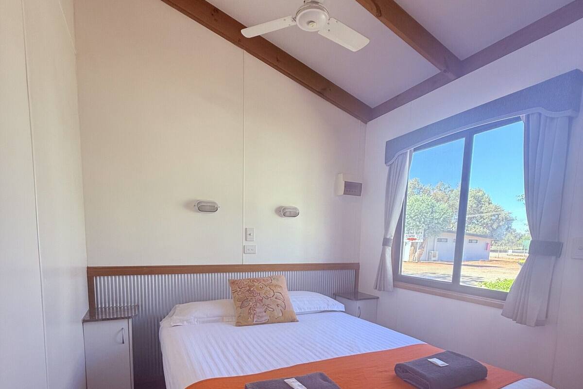 Habitación de hotel con cama, mesita de noche, ventilador de techo, ventana con cortinas y vistas al exterior.
