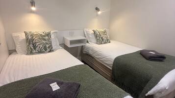 A hotel room with two single beds, each with a green blanket and a white pillow. There are wall-mounted lights above each bed.