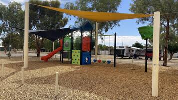 Children can play safely on the playground's soft surface, surrounded by trees and featuring a slide, climbing panels, and swings.