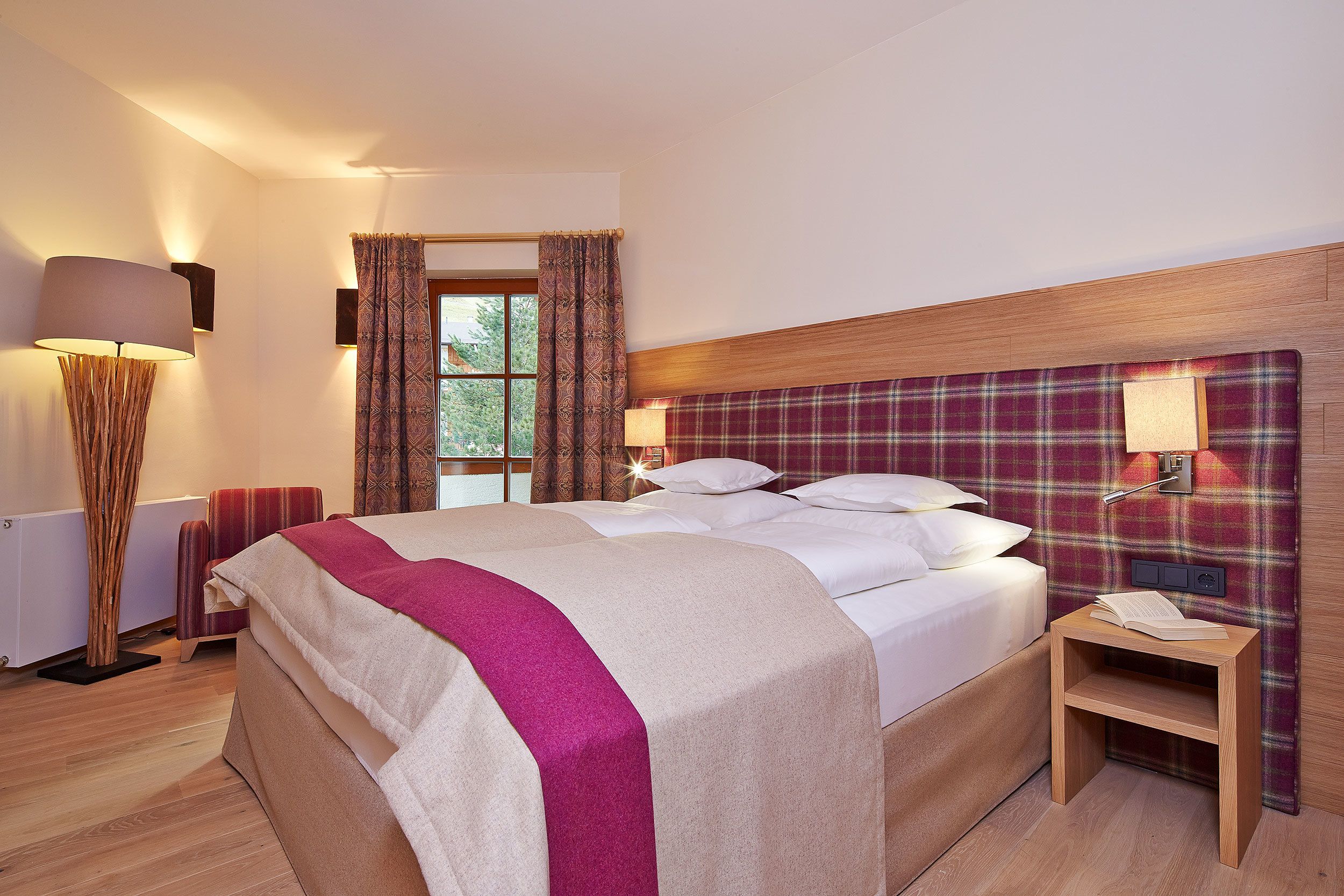 A neatly made bed with a plaid headboard, a bedside table with a lamp, and a window with curtains.