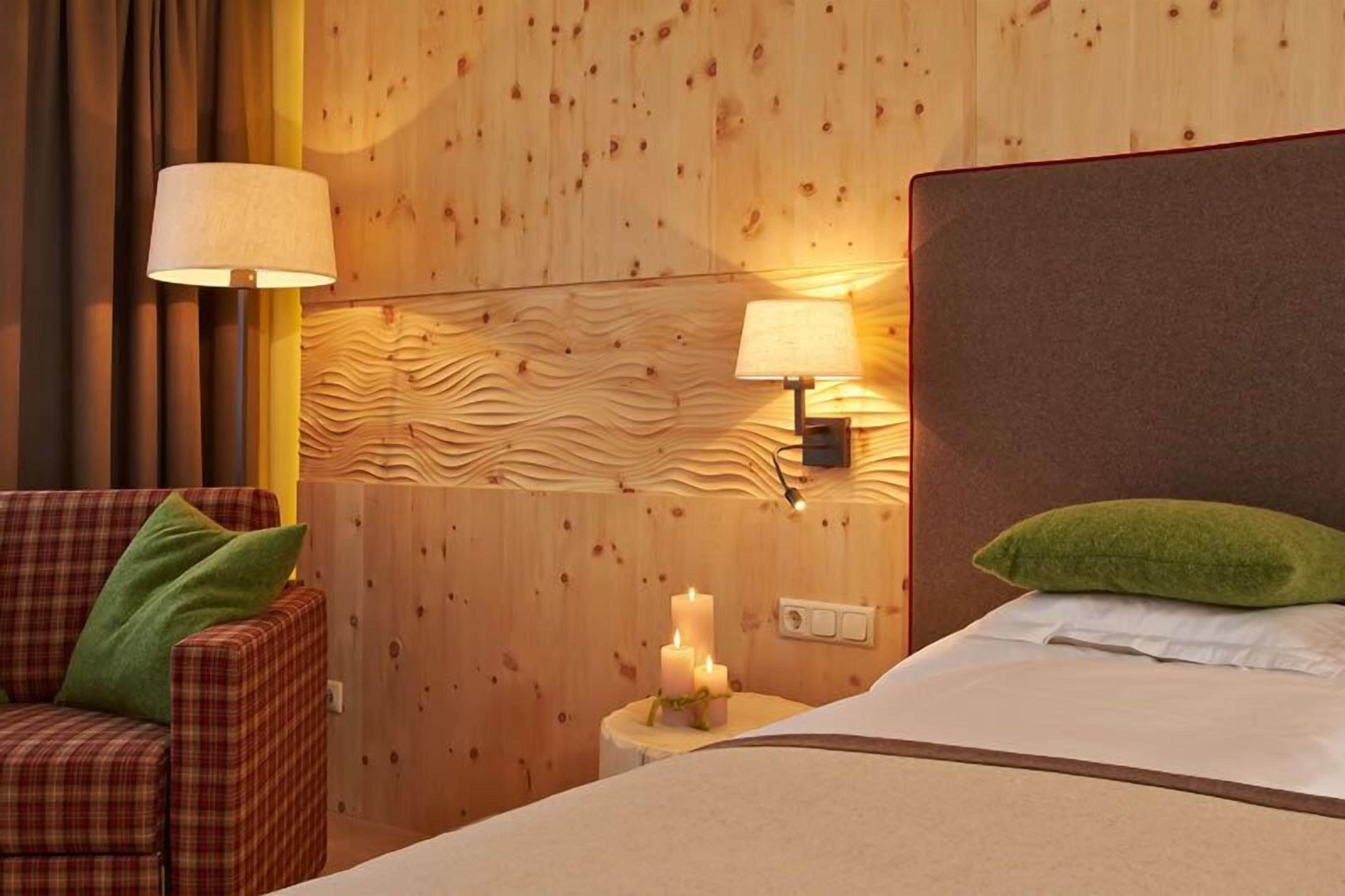 A hotel room with a wooden panel wall, a bed with white bedding, a plaid chair with green cushions, two wall-mounted lamps, and a small table with candles.
