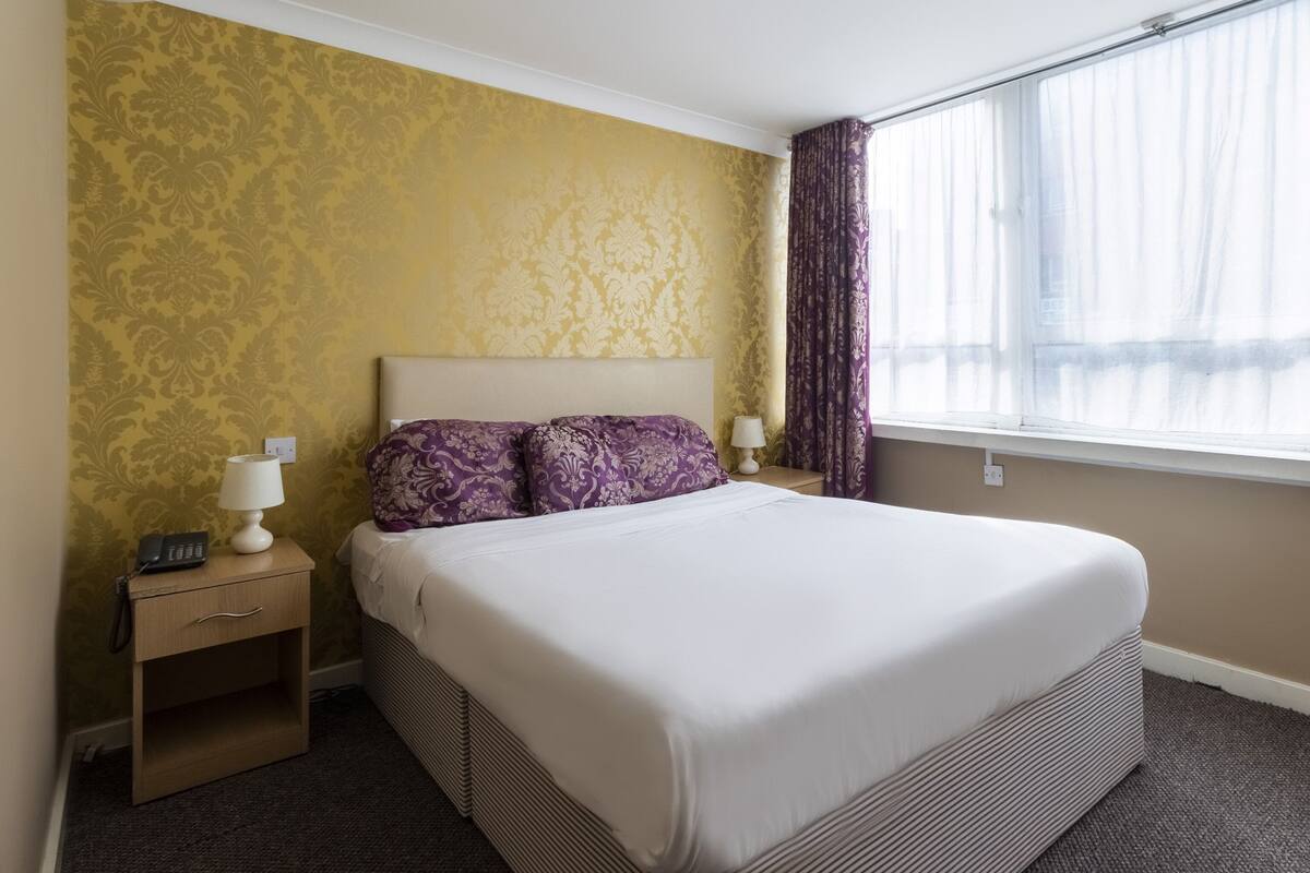A hotel room with a large bed, a bedside table with a lamp, a phone, and a nightstand.