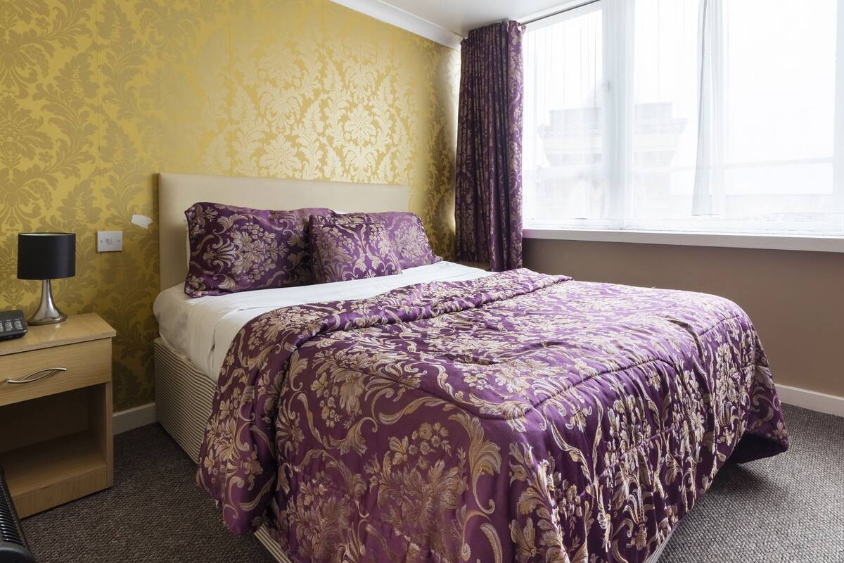 A hotel room with a bed, a nightstand, a lamp, and a window with curtains.