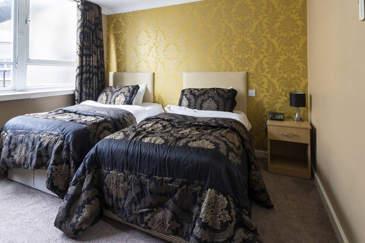 A hotel room with two beds, a bedside table with a lamp, and a window with curtains.