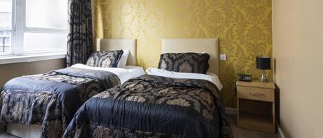 Una habitación de hotel con dos camas, una mesita de noche con lámpara y una ventana con cortinas.