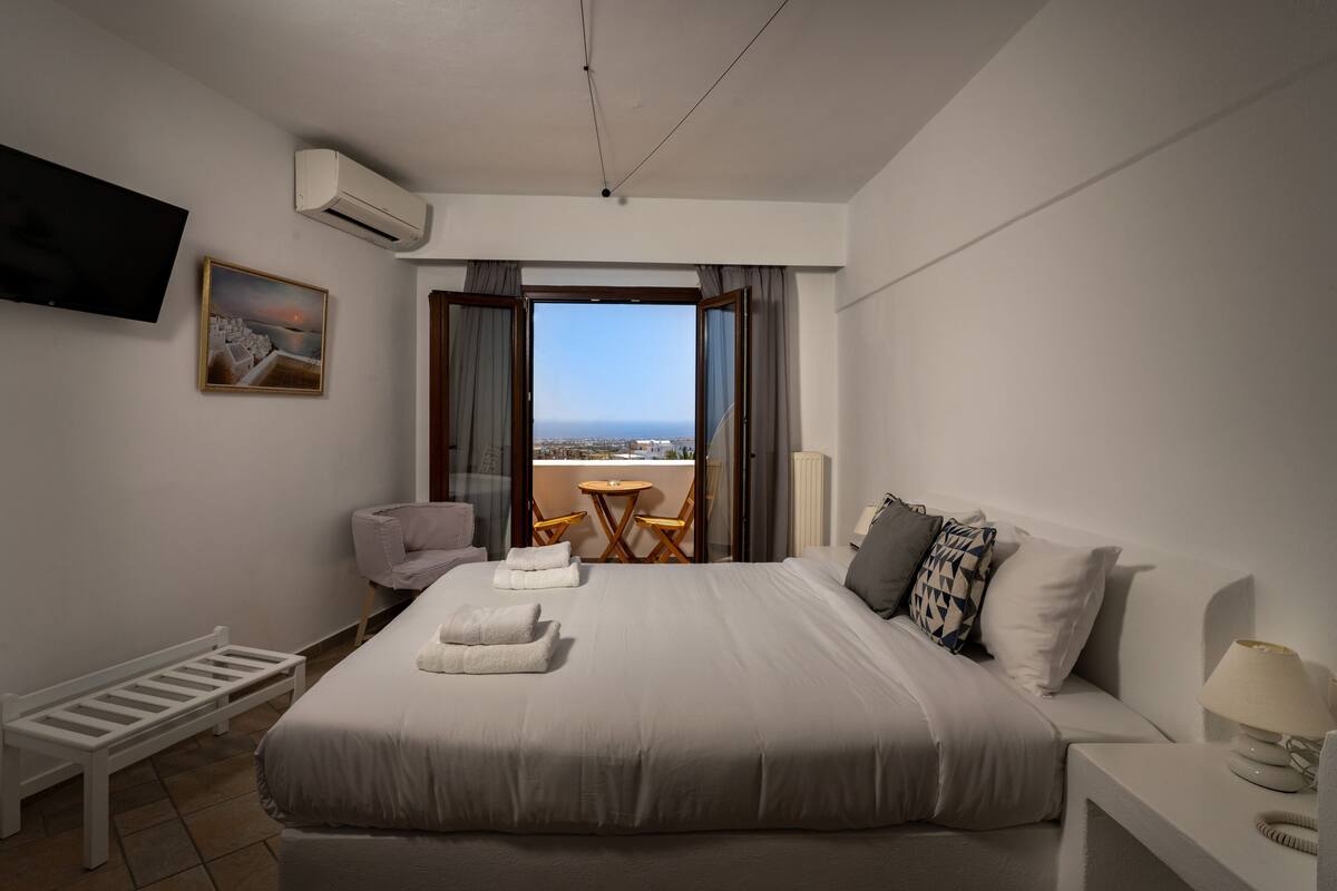 A hotel room with a large bed, a TV, a painting, a balcony with a view, and a small table with chairs.