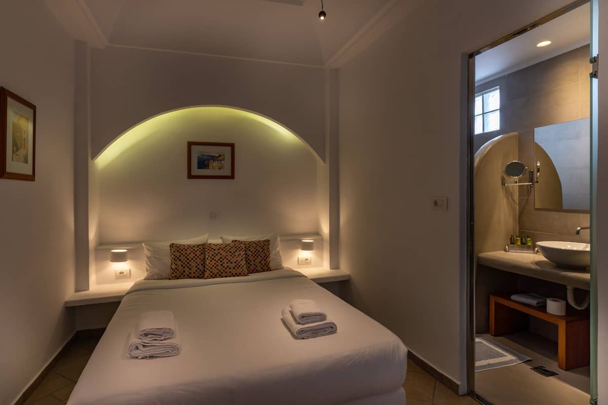 A hotel room with a neatly made bed, a bedside table with a lamp, a large mirror, and a bathroom with a sink and mirror.
