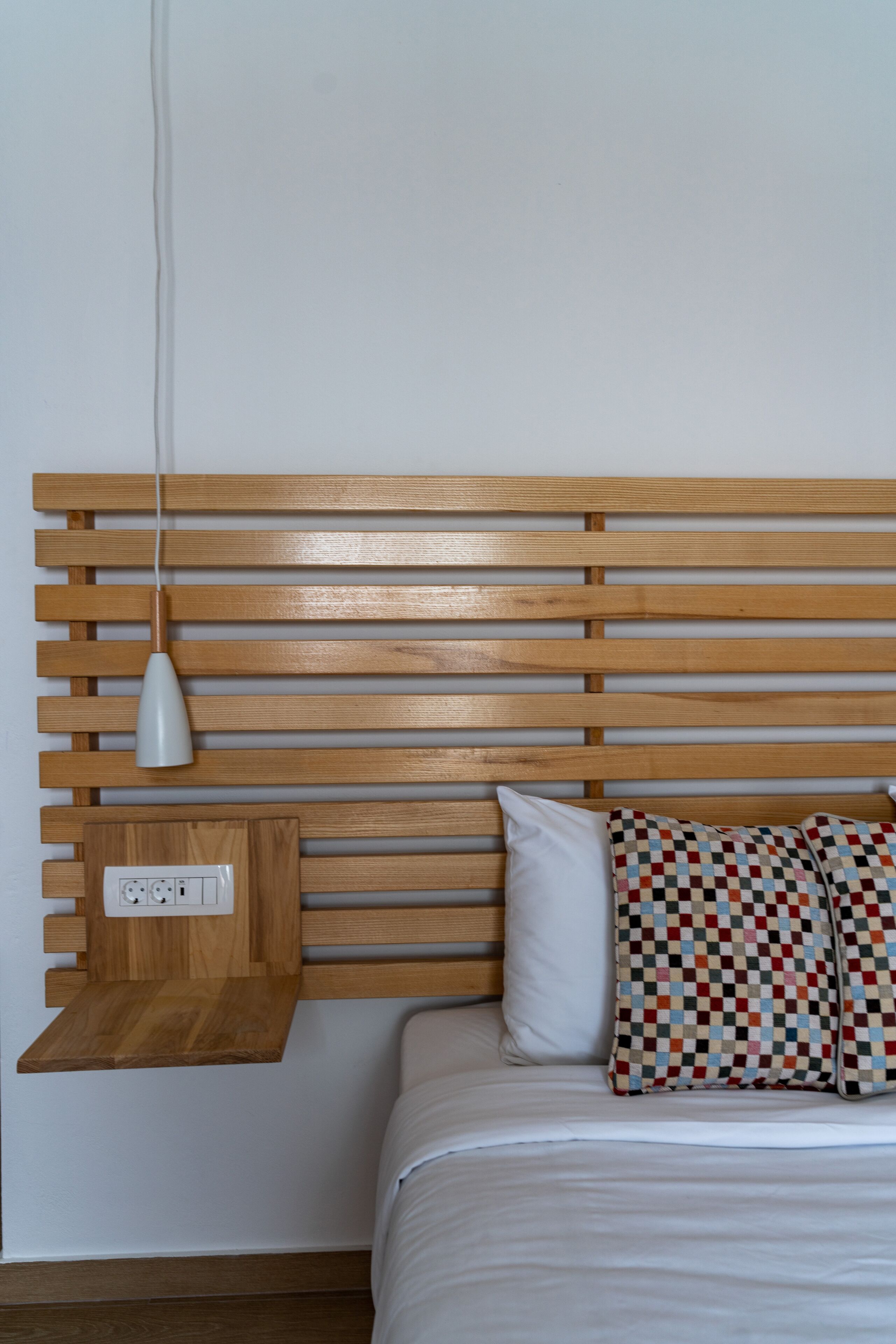 Une chambre à coucher avec une tête de lit en lattes de bois, une table de chevet dotée d’une prise électrique, un oreiller blanc et un coussin décoratif à motifs.