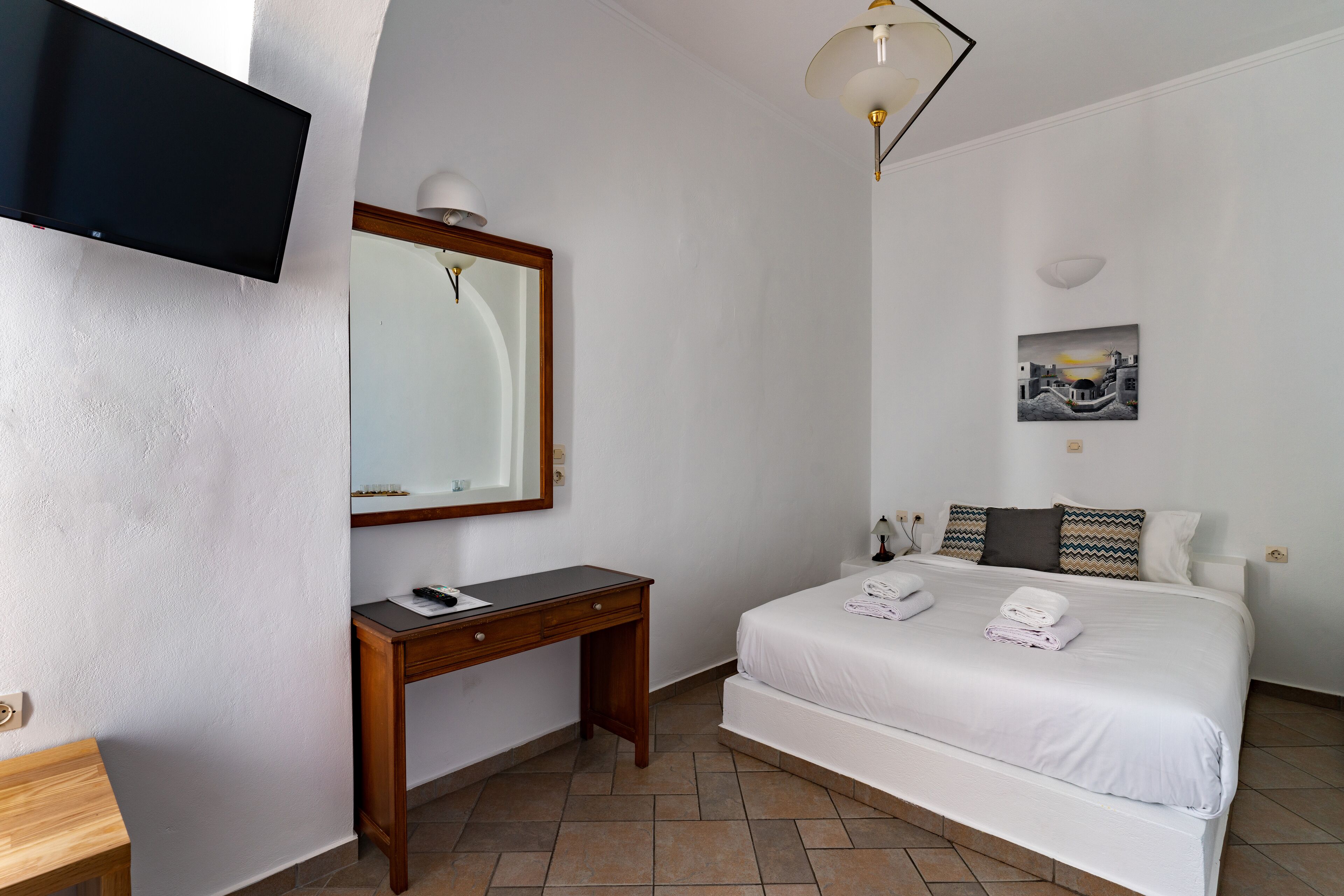A hotel room with a bed, a desk, a flat-screen TV, a mirror, a wall-mounted lamp, and a painting.